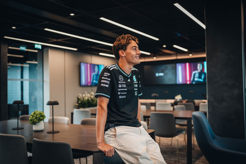 George Russell, driver for the Mercedes-AMG PETRONAS Formula One Team, is casually sitting on a table in a modern, stylish team hospitality or office area. He’s smiling and wearing the official team shirt with sponsor logos such as Einhell, IWC, and CrowdStrike visible. Screens in the background show his image.