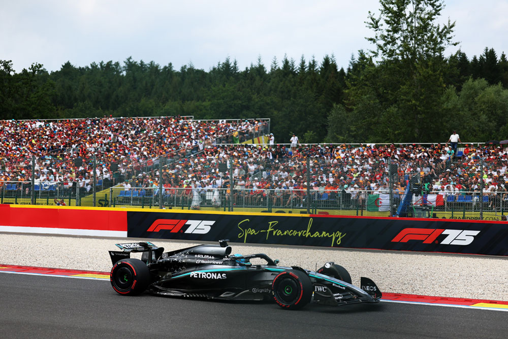 A Mercedes-AMG PETRONAS Formula One car is racing on the Spa-Francorchamps circuit. In the background, packed grandstands are filled with fans waving international flags. The Spa-Francorchamps logo is displayed on the barrier.