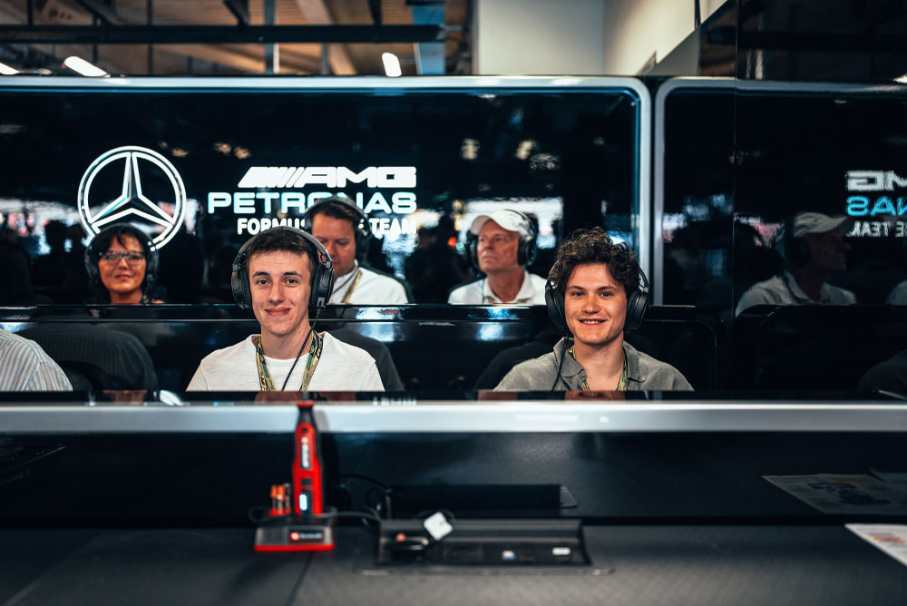 Two men are sitting in the Mercedes-AMG PETRONAS Formula One Team garage, wearing headsets and enjoying the behind-the-scenes experience. A red Einhell device is visible in the foreground.
